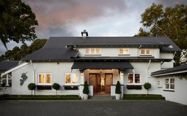 Front exterior view of a substantial two-storey residence with a symmetrical light-rendered facade, steep dark metal gabled roof, covered entrance porch, timber double front doors, dormer-like upper windows, white-framed windows, and neatly clipped hedges and topiary. The home has a refined traditional character with modernised detailing, photographed in soft overcast evening light.