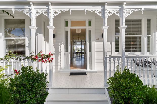 Front exterior of a refined white weatherboard home with a raised porch, decorative lacework brackets and fretwork, turned timber posts and balustrades, white-framed windows with shutters, and a central glazed entry door. The symmetrical facade is surrounded by manicured shrubs and flowering plants, creating a bright heritage-style streetscape.