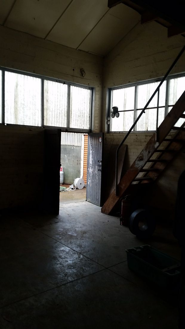 A dark, utilitarian industrial interior with exposed masonry walls, a concrete floor, high translucent strip windows, an open metal door to the exterior, and a rough timber-and-metal staircase leading upward. Workshop or warehouse equipment is visible in shadow.