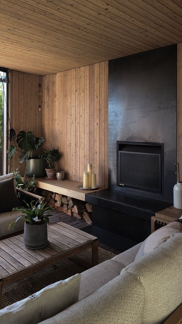 A warm, intimate living room with extensive vertical timber wall panelling and a matching timber-lined ceiling. A dark fireplace surround and built-in black fireplace form a strong feature wall, with a timber hearth bench and neatly stacked firewood beneath. The room contains upholstered sofas, a timber coffee table, potted plants, candles, and textured neutral furnishings. The partially glazed side opening brings in natural light and greenery, creating a cozy indoor-outdoor atmosphere.