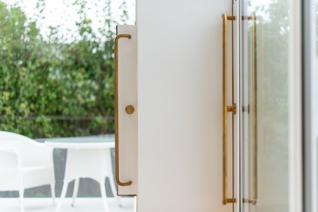 Close-up architectural detail of a glazed exterior door with large brass-toned vertical pull handles and a white frame, opening onto a bright outdoor terrace or patio with white furniture and leafy greenery beyond. The image focuses on the door hardware, glazing, and threshold rather than a room interior.