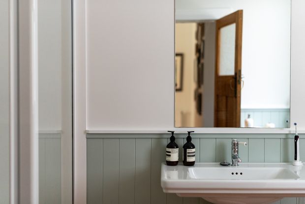 A compact, traditional-style bathroom vanity area with a white wall-mounted ceramic basin, chrome tapware, pale sage-green vertical wall panelling, and white painted trim. A large mirror reflects an open timber door and part of the adjoining interior. Dark soap dispensers and a toothbrush add small functional details.