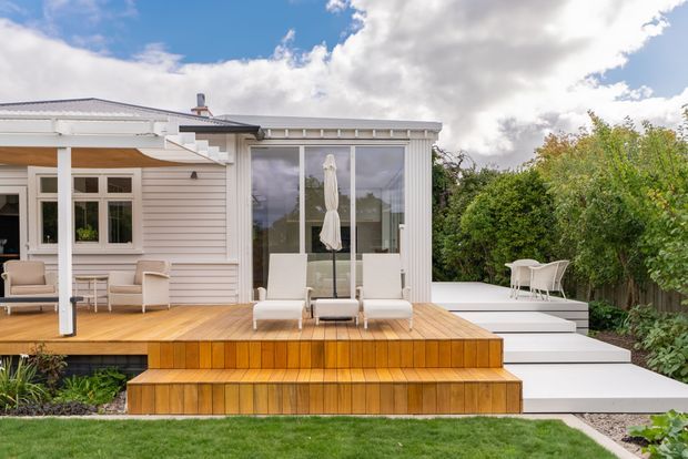 Bright exterior view of a light-coloured weatherboard home opening onto a broad timber deck with stepped built-in seating or stairs. Large glazed doors, white-framed windows, outdoor lounge furniture, and a white-paved side terrace are surrounded by established garden planting. The home has a low-pitched roof and a clean, relaxed outdoor entertaining setup.