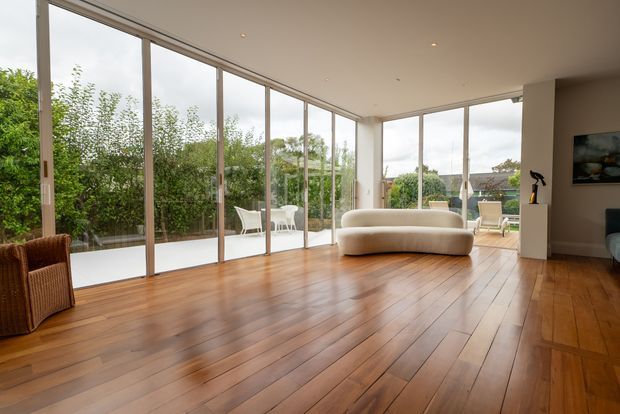 A bright, spacious contemporary living room with warm timber flooring, a curved cream sofa, and extensive floor-to-ceiling sliding glass doors opening toward a landscaped outdoor area. Minimal recessed lighting, slim window frames, and a restrained neutral palette create an airy indoor-outdoor connection.