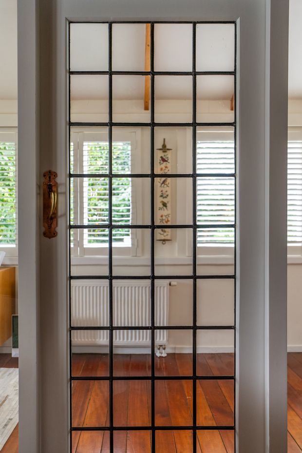 A tall, dark-framed multi-pane glass door or partition with a warm timber handle, reflecting and revealing a bright room beyond. The room has white plantation shutters, a traditional white radiator, pale walls, and polished reddish-brown timber flooring. The image focuses on the glazed architectural divider and its reflections.