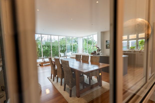A refined open-plan dining area viewed through a glazed doorway or partition. A large dark timber dining table with woven upholstered chairs sits on a neutral rug over polished timber flooring. Floor-to-ceiling glazing wraps around the bright room, opening to leafy greenery, while a kitchen or serving area is visible to the right with dark cabinetry and a large rounded pendant light.