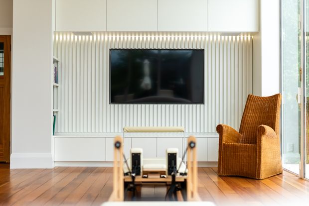 Bright contemporary living room with a wall-mounted television set into a white built-in media wall featuring vertical fluted detailing and integrated strip lighting. The room has warm timber flooring, a woven tan armchair, floor-to-ceiling glazing at right, and a partially visible exercise machine in the foreground.