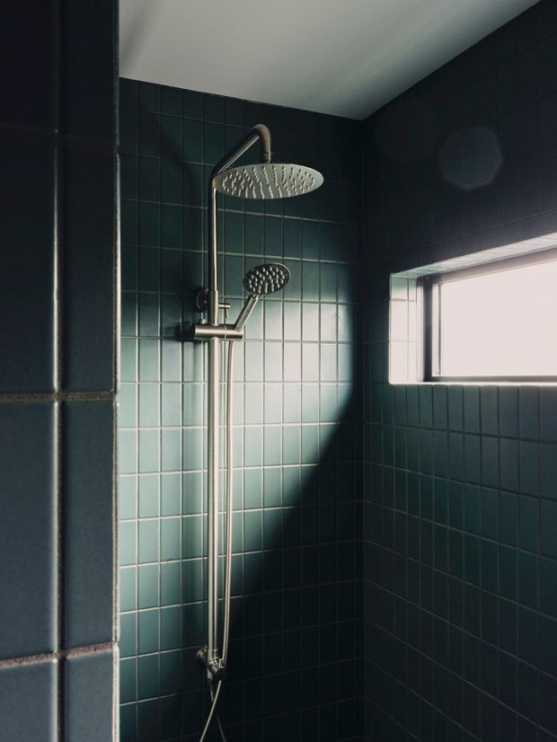 A compact, moody shower enclosure with dark teal square wall tiles, a brushed metal dual shower system, and a small horizontal window admitting strong natural light. The dramatic light and shadow create a refined, atmospheric bathroom setting.
