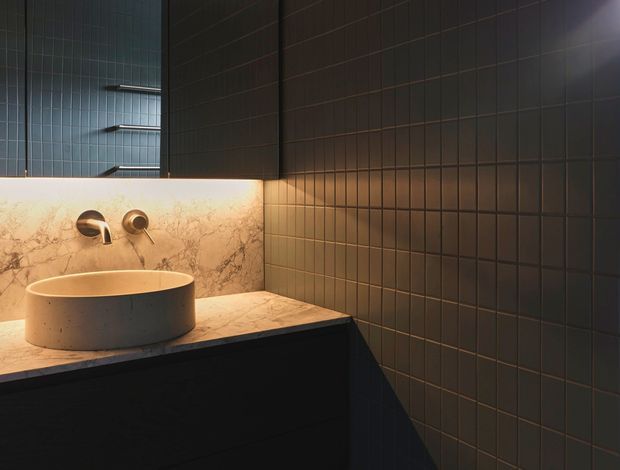 A moody contemporary bathroom vanity featuring a cylindrical concrete vessel basin on a pale veined stone benchtop. Dark rectangular wall tiles wrap the corner, with matching dark cabinetry below, brushed metal wall-mounted tapware, and warm concealed lighting behind the stone splashback.