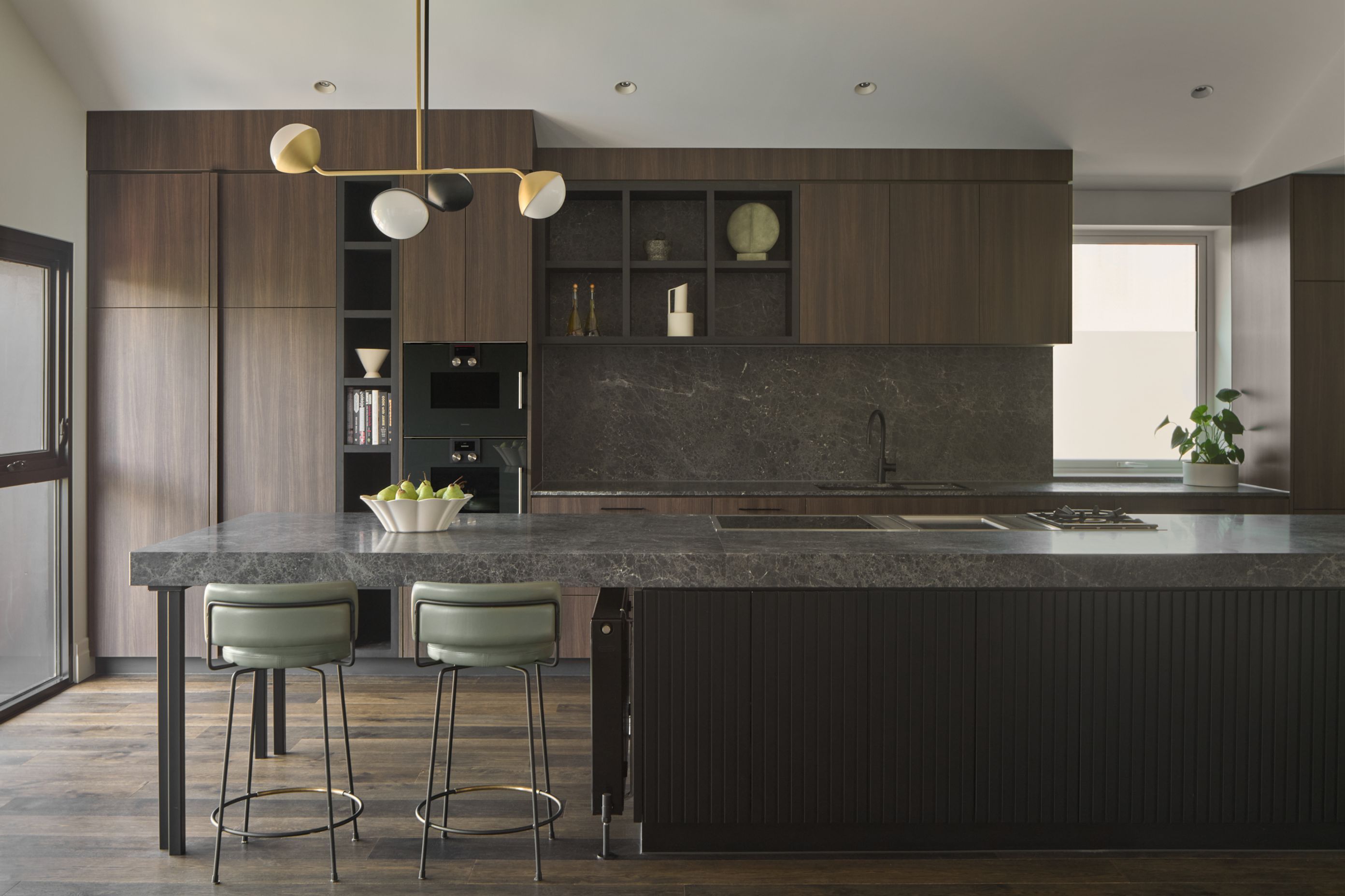 At the heart of the home, the kitchen is a study in both form and function. An expansive island anchors the space, with custom seating and pendant lighting adding sculptural flair. Art Deco references shine through—tailored, warm, and wonderfully livable.