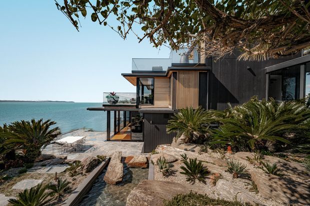 A modern multi-level coastal house set directly beside turquoise water, viewed from a landscaped rocky garden. The building combines dark vertical cladding, warm timber detailing, extensive glazing, projecting balconies, and a flat roofline. Succulents and cycads surround stone and concrete pathways leading toward the waterfront, while outdoor seating and a lower-level terrace are visible.