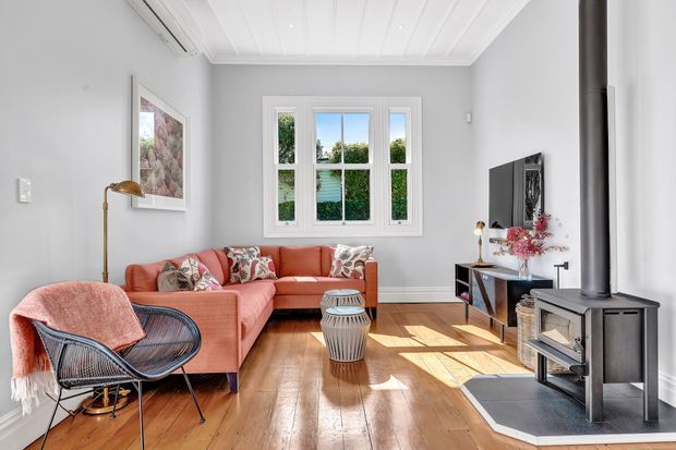 A bright, warmly furnished living room with a coral sectional sofa, woven lounge chair, timber flooring, white paneled ceiling, grey walls, and a central wood-burning stove on a dark hearth. A large multi-pane window brings in abundant daylight, while a wall-mounted television, console, artwork, and floral decor complete the room.
