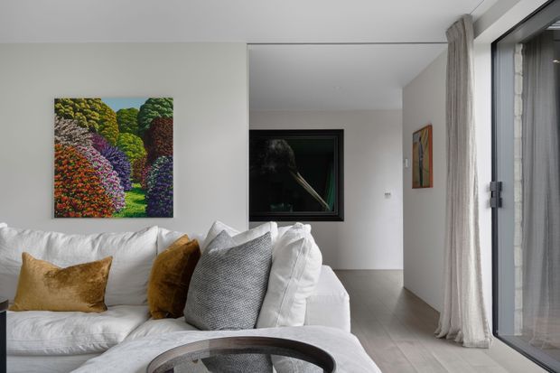 Bright contemporary living room with a large white sectional sofa, textured grey and mustard cushions, light grey flooring, white walls, full-height glazed doors or windows with sheer curtains, and colourful artwork. A hallway or adjoining space is visible beyond the seating area.