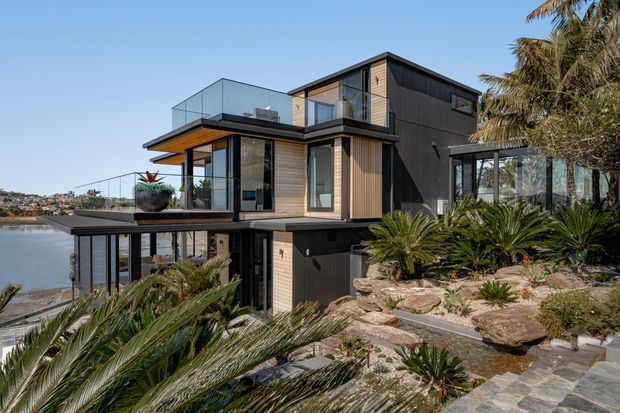 Modern multi-level residence on a sloping waterfront site, featuring dark cladding, warm timber-look wall panels, extensive glazing, projecting terraces with glass balustrades, and a landscaped rocky garden with cycads. The home overlooks calm water and a developed shoreline in the distance.
