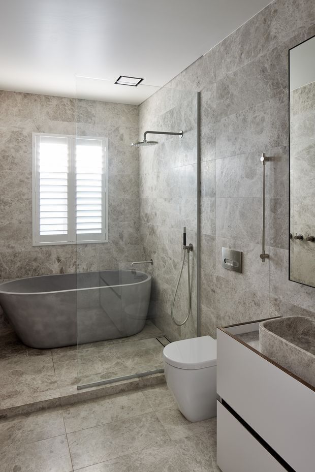 A refined contemporary bathroom featuring full-height grey stone-look wall and floor tiles, a freestanding grey bathtub beneath a shuttered window, a frameless glass shower enclosure with brushed metal fixtures, a wall-hung toilet, and a minimalist white floating vanity with a stone vessel basin. A large mirror and recessed ceiling vent complete the clean, neutral scheme.
