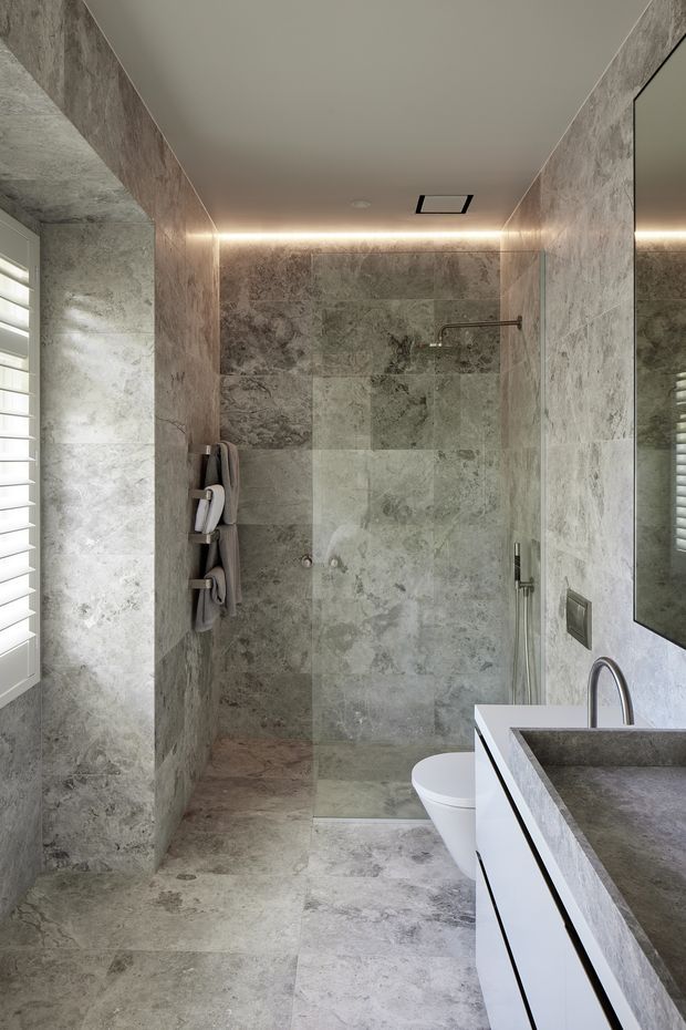 Contemporary bathroom with a walk-in tiled shower, wall-mounted white toilet, floating white vanity with a grey stone benchtop, large mirror, and grey natural-stone-look wall and floor tiles. Integrated linear lighting and a louvred window create a refined, spa-like atmosphere.