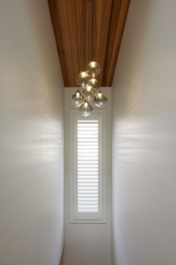 A narrow, tall hallway or stairwell with smooth white walls, a warm timber-lined ceiling, and a cluster of hanging glass pendant lights. A slim vertical window with white plantation shutters is centered at the far end, creating a bright, refined architectural feature.