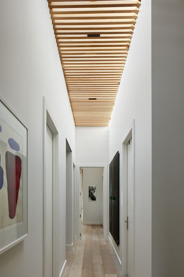 A narrow, bright residential hallway with white walls, pale timber flooring, multiple doorways, and a striking open timber slat ceiling creating a linear architectural feature. Framed abstract artwork is visible along the left wall, with additional artwork at the far end.