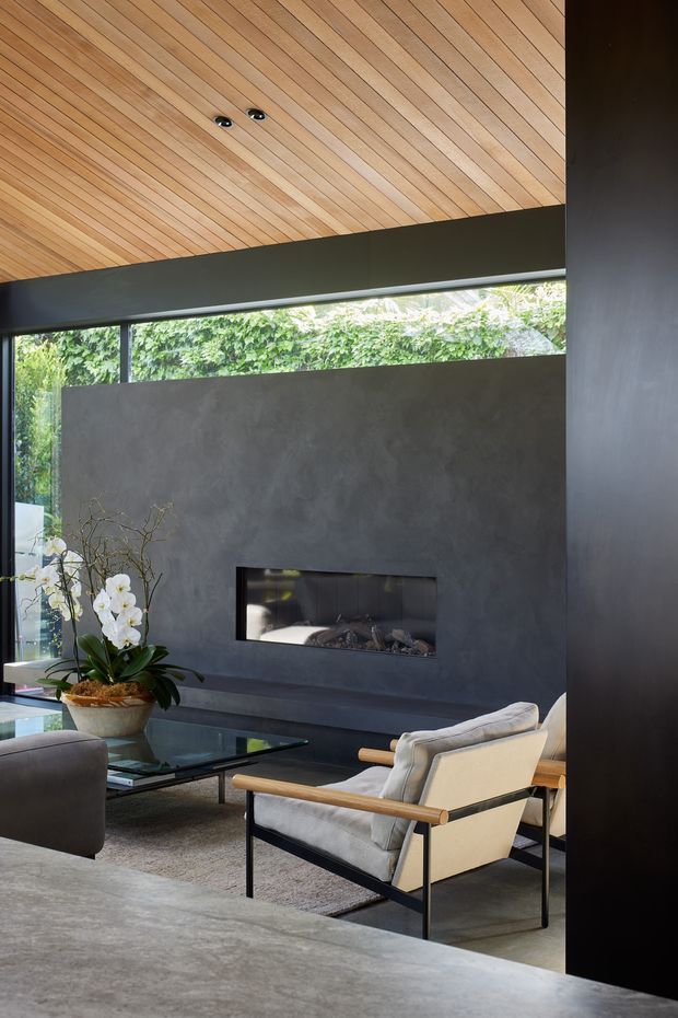 A sophisticated living room with a low-profile fireplace set into a dark textured feature wall, floor-to-ceiling glazing, and a warm timber-lined ceiling. The room includes contemporary lounge chairs, a glass coffee table, a large neutral rug, and an indoor orchid. Dark structural framing contrasts with the natural timber ceiling and lush greenery visible outside.