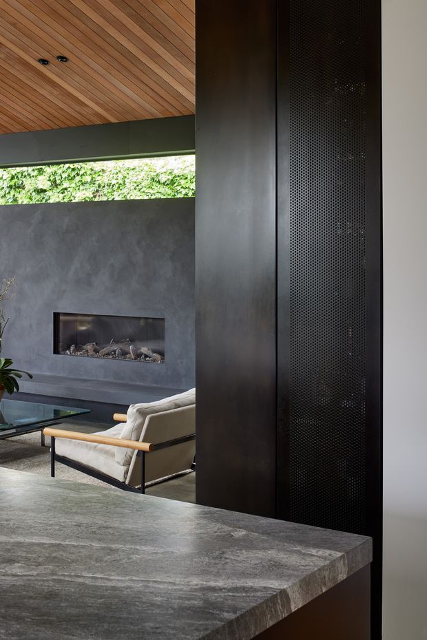 A refined open-plan living area viewed from behind a dark stone kitchen or counter. The room features a dramatic charcoal textured fireplace wall with a recessed linear fire, a black perforated metal screen or partition, a pale upholstered lounge chair with timber armrests, a glass coffee table, and a warm slatted timber ceiling. A narrow clerestory opening brings in greenery and daylight.