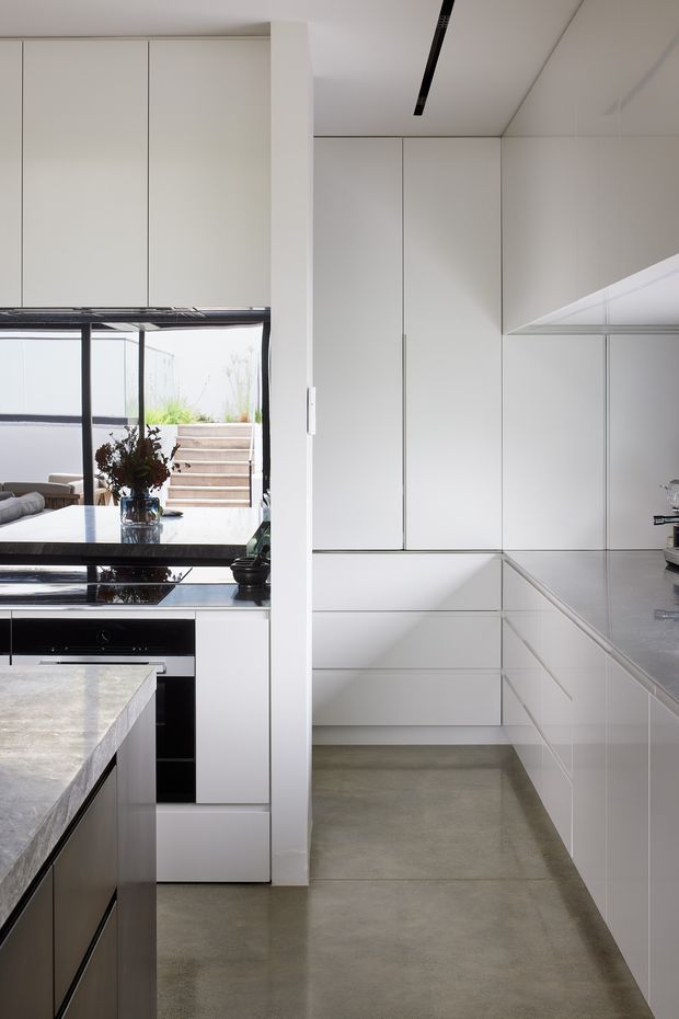 A bright, minimalist contemporary kitchen with extensive floor-to-ceiling white cabinetry, integrated appliances, a large stone island and benchtops, and a polished concrete floor. Black-framed glazing opens to an outdoor stair and planted area, while a linear black ceiling light adds contrast.
