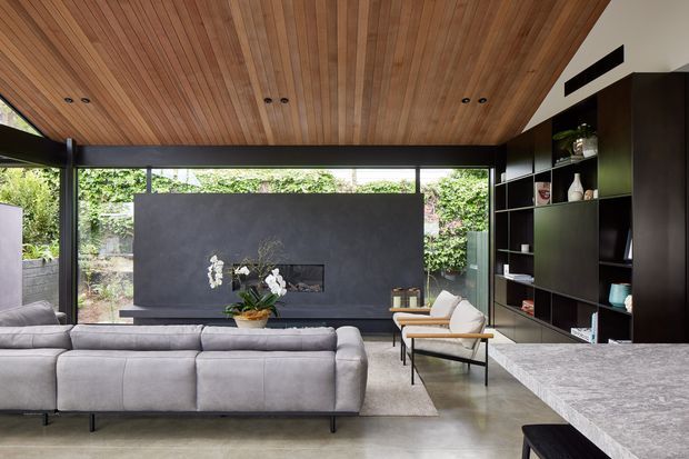 Contemporary living room with a large grey sectional sofa facing a dark feature fireplace wall, expansive black-framed glazing opening to greenery, a warm timber-lined ceiling, and full-height dark built-in shelving. A stone kitchen island occupies the foreground, suggesting an open-plan living and kitchen area.