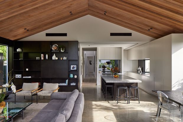 Open-plan contemporary living, dining and kitchen interior with a dramatic pitched timber-lined ceiling. The room features dark built-in shelving, a large grey sectional sofa, lounge chairs, a stone kitchen island with bar seating, pale cabinetry, polished concrete flooring, and strong indoor-outdoor light.