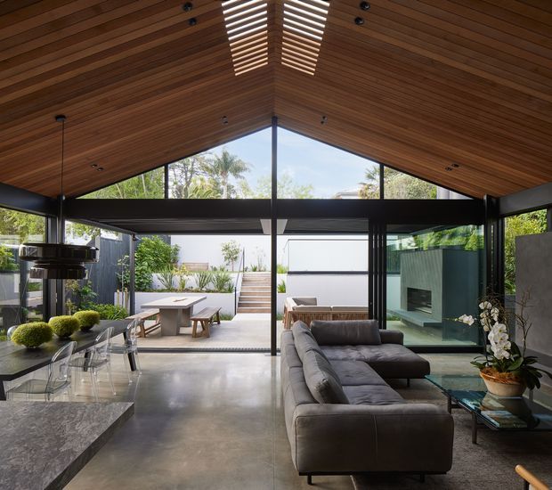 Open-plan contemporary living and dining space with a dramatic pitched timber-lined ceiling and exposed dark structural framing. Full-height glazing opens onto a landscaped courtyard, while a large sectional sofa faces a wall-mounted fireplace and a polished stone kitchen island anchors the foreground. The room has polished concrete flooring, abundant natural light, and a strong indoor-outdoor connection.