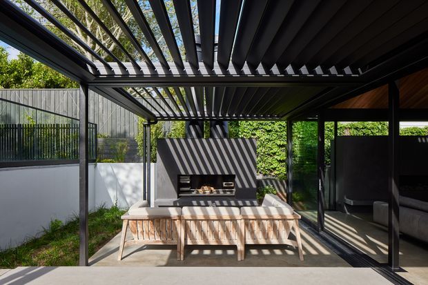 A contemporary covered outdoor entertaining area with a dark steel pergola featuring adjustable louvres, a built-in barbecue set into a rendered wall, and timber outdoor dining furniture. Glazed sliding doors connect the space to an adjoining interior, while a private landscaped courtyard and fencing surround the area. Strong patterned sunlight and shadows create a crisp, modern atmosphere.