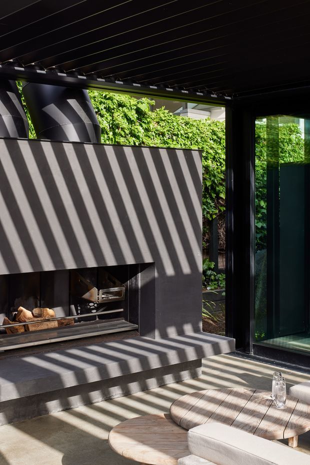 A contemporary outdoor living area beneath a dark slatted pergola, featuring a built-in concrete fireplace with a wood-burning firebox, black-framed glazing, and lush greenery beyond. Strong linear shadows fall across the fireplace, paving, and rounded timber outdoor coffee tables, creating a dramatic yet inviting atmosphere.
