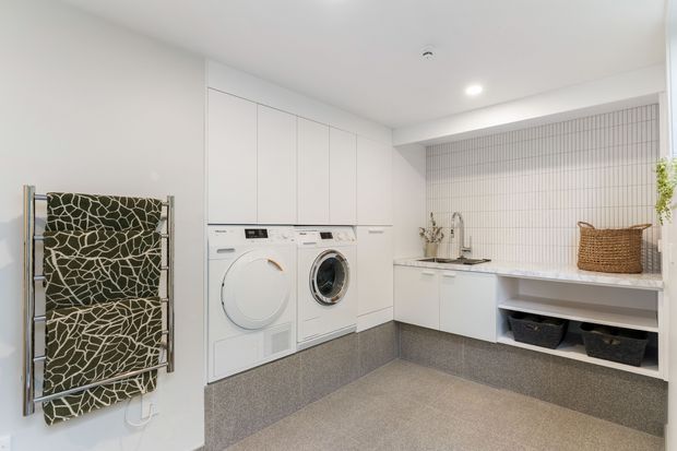 Bright, contemporary laundry room with white full-height cabinetry, integrated front-loading washer and dryer, a long white benchtop, stainless-steel sink and tap, and a recessed open shelving area. The room features a grey small-format tiled splashback, grey tiled floor and matching grey tiled cabinetry plinth, with a patterned heated towel rail and woven basket adding visual interest.