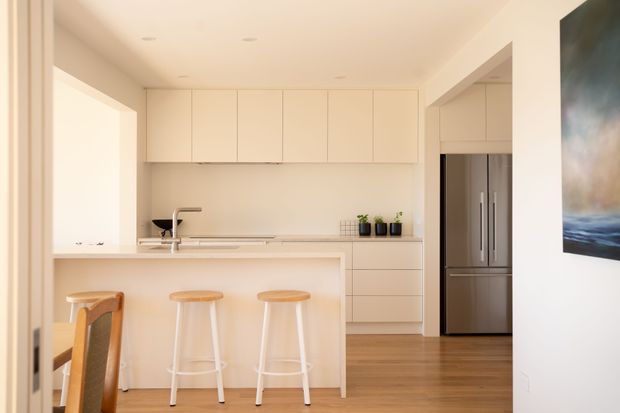 A bright, minimalist kitchen with a large pale island and three timber-topped stools, flat-panel cream cabinetry, integrated appliances, stainless-steel refrigerator, timber flooring, and recessed ceiling lights. A dining chair and framed artwork are visible at the edges of the image.