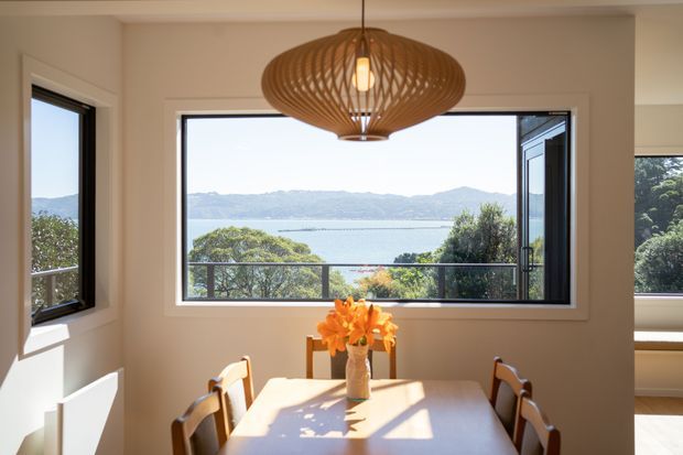 Bright dining room with a rectangular timber dining table and chairs, large black-framed windows, and a sculptural woven pendant light. The windows overlook a calm bay or waterfront landscape, while warm neutral walls and abundant daylight create an inviting atmosphere.