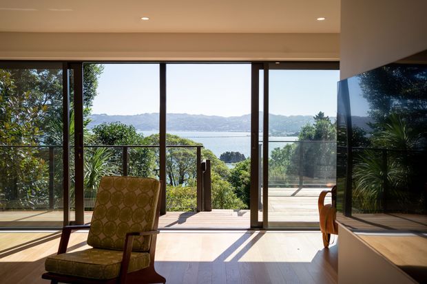 A bright contemporary living room opening onto a timber deck, with full-height glazed sliding doors framing a leafy garden and expansive water and mountain views. The room has warm timber flooring, recessed ceiling lights, a patterned upholstered lounge chair, and a large reflective dark surface or television wall on the right.