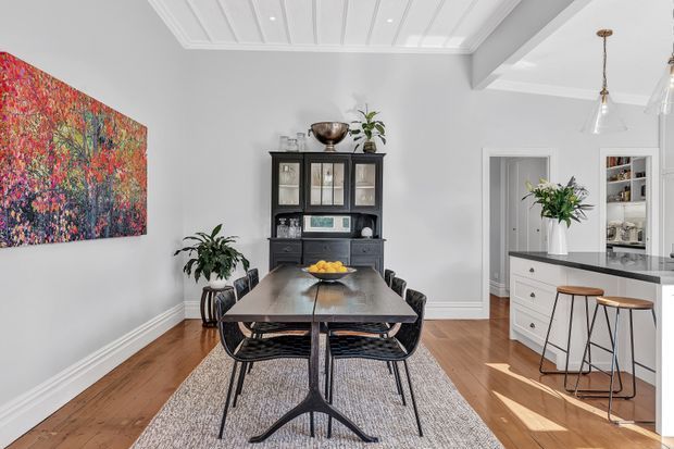 Bright, refined dining area in an open-plan home, featuring a dark timber dining table with black-framed chairs, a large textured artwork, a black built-in or freestanding display hutch, warm timber flooring, and a white kitchen with island seating visible to the right. The room has white walls, decorative panelled ceiling detailing, pendant lighting, and abundant natural light.