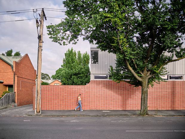 Street-level view of a contemporary urban building behind a long red brick boundary wall. The facade combines horizontal red brickwork with pale grey vertical metal cladding and narrow horizontal windows. A mature street tree, utility pole, overhead wires, sidewalk, and residential brick buildings frame the scene.
