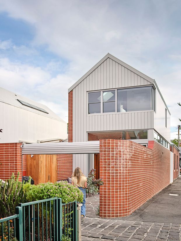 Street-level exterior view of a contemporary multi-storey residence with a steep gabled roof, pale vertical cladding, extensive upper-level glazing, and prominent red brick walls. A curved brick boundary wall encloses a small landscaped front courtyard, with a timber gate, covered entry structure, paved walkway, and neighbouring urban buildings visible.