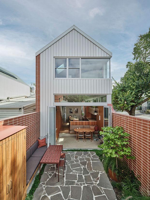 A narrow urban courtyard viewed toward a compact multi-level house. The facade combines vertical light-grey metal cladding, red brick side walls, a steep gabled roof, and extensive glazing. The ground level opens onto a paved courtyard with a built-in dark bench, outdoor dining table and chairs, timber storage cabinetry, brick boundary walls, and planted greenery. Warm timber interiors and furnishings are visible through the open glazed doors.