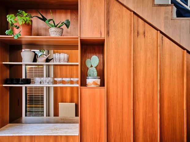 Built-in timber cabinetry and shelving integrated beneath and alongside a staircase. Warm wood joinery features open display shelves with plants, ceramics, and glassware, plus a pale stone or marble-look benchtop. The stair enclosure has vertical timber panels and a dark handrail, creating a strongly crafted mid-century-inspired interior feature.