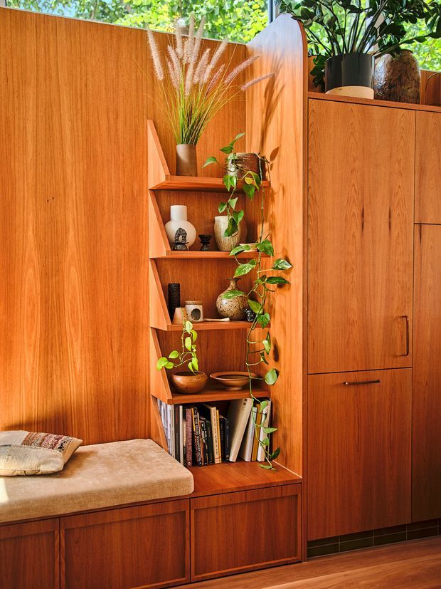 Warm, richly detailed built-in timber joinery forms a compact shelving nook beside a tall storage cabinet. The shelving is styled with books, ceramic vessels, potted trailing plants and dried grasses, while a cushioned built-in bench occupies the lower left. Matching wood panelling continues across the wall and cabinetry, creating a cohesive mid-century-inspired interior vignette.