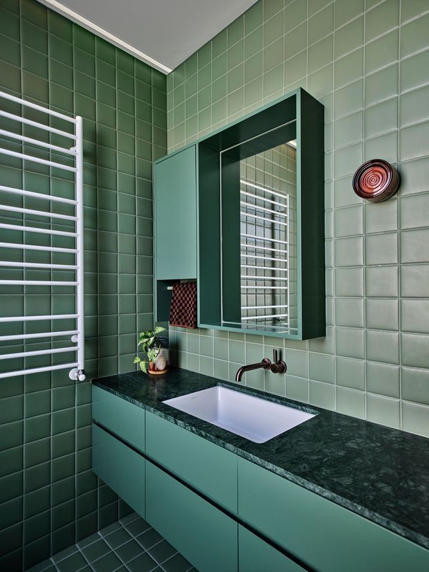 A compact, highly coordinated green bathroom featuring full-height square green wall tiles, matching floor tiles, a dark green stone benchtop, and teal-green vanity cabinetry. A white inset basin sits beneath a large framed mirrored cabinet, with a wall-mounted bronze tap and white heated towel rail. The room has a bold, refined retro-modern character with strong tonal colour continuity.