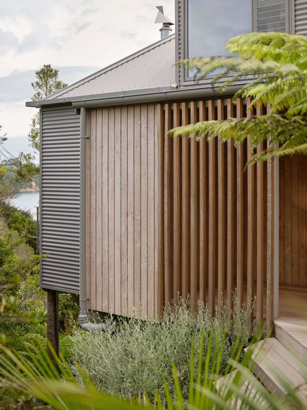 Exterior view of a compact elevated home with a steep corrugated metal gable roof, grey ribbed metal cladding, pale vertical timber weatherboards, and a screened timber entry porch. A small roof vent and louvred upper window are visible, with landscaped planting in the foreground and steps leading to the entrance.