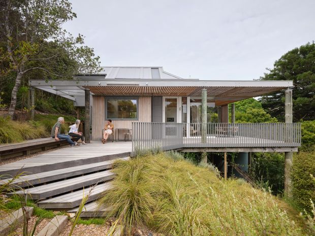 Exterior view of a contemporary elevated pavilion-style house set into a landscaped, sloping site. The building has a low-pitched metal roof, corrugated or ribbed grey wall cladding, extensive covered outdoor decking, timber pergola slats, and a grey balustrade. Broad timber steps lead up through native grasses and vegetation to the deck, with people seated outside.