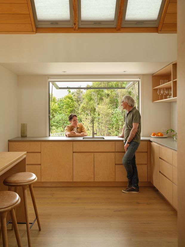Warm, compact contemporary kitchen with extensive light timber cabinetry, a peninsula with stools, a large open awning window above the sink, and clerestory-style roof windows. The kitchen opens visually to dense greenery outside and includes open shelving with glassware and a stone-look benchtop. Two people are present but the focus is the fitted kitchen interior.