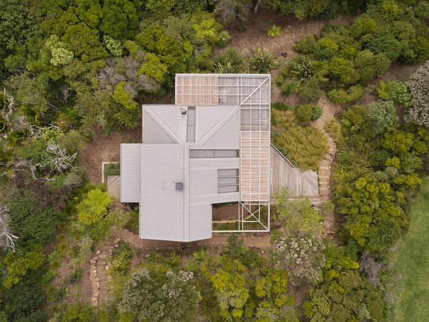 Top-down aerial view of a contemporary residential building nestled within dense native vegetation. The house has multiple rectilinear roof volumes in pale grey metal, a prominent screened or framed rooftop terrace/structure, and timber-toned deck or walkway elements. Winding stone paths connect the building through the landscaped bush setting.