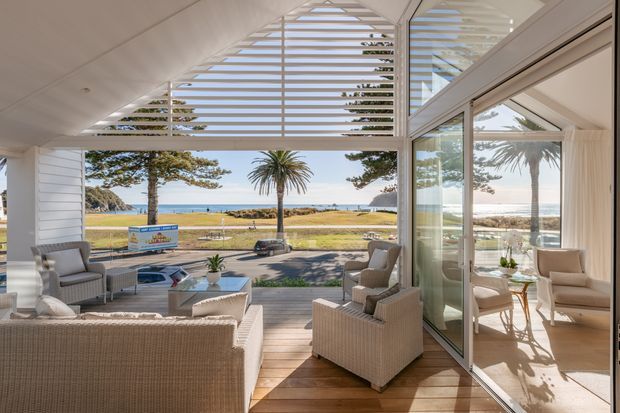 Bright coastal living area opening onto a timber deck, furnished with woven lounge seating and low tables. Large glazed sliding doors and a triangular louvred clerestory frame views toward a sandy beach, ocean, and landscaped coastal reserve. The reflected interior shows additional seating and abundant natural light.
