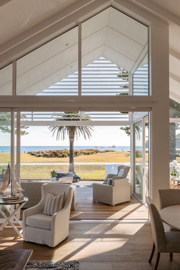 Bright coastal living and dining area opening onto a timber deck, with expansive floor-to-ceiling glazing, white-framed windows, and a slatted triangular clerestory beneath a pitched ceiling. The room is furnished with light upholstered armchairs, wicker outdoor seating, and a dining table. A palm-lined lawn and ocean view are visible beyond the open doors.
