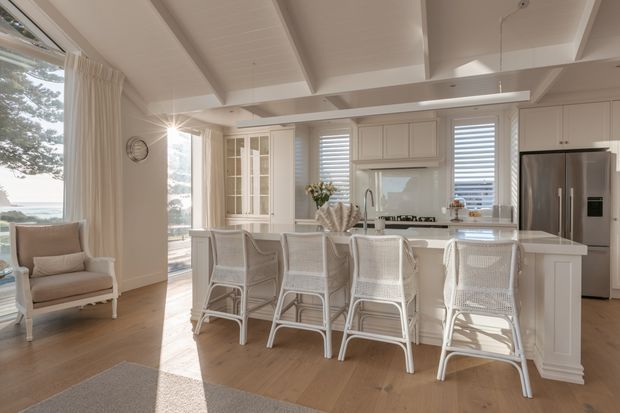 Bright coastal-style kitchen and dining space with a large white island, four woven-back bar stools, white shaker cabinetry, stainless-steel refrigerator, plantation shutters, and a white painted timber ceiling with exposed beams. Large windows and glazed doors bring in abundant natural light, with a lounge chair positioned by the window.