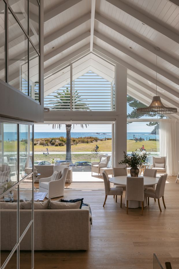 Bright coastal open-plan interior with a living and dining area facing the ocean. The room has a dramatic double-height pitched ceiling lined with white timber boards, exposed white beams, extensive glazing and a large central gable window with horizontal louvres. Pale timber flooring, neutral upholstered seating, a round dining table, woven pendant light and indoor greenery create a relaxed seaside atmosphere. A beach, lawn and ocean are visible through the glazing.