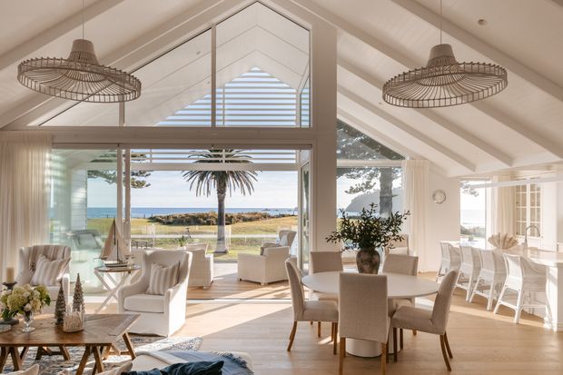 Bright, open-plan coastal living and dining area with a vaulted ceiling and prominent triangular clerestory glazing. The room features pale timber flooring, white walls and furnishings, woven pendant lights, a dining table, and a relaxed seating area. Large glazed doors open toward a lawn, palm tree, and ocean horizon.